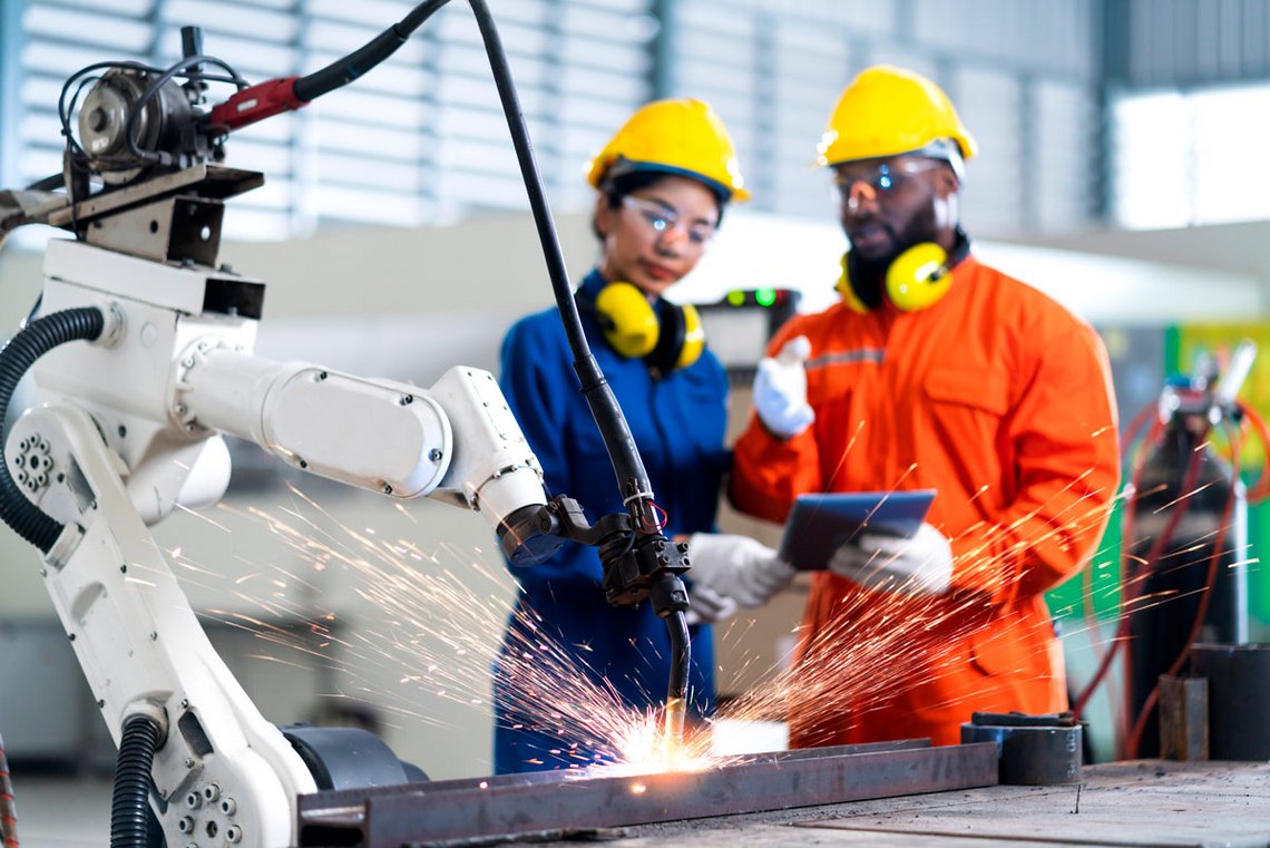 engineer cooperation male and female technician maintenance control relay robot arm system welding with tablet laptop to control quality operate process work heavy industry 4.0 manufacturing factory Der Cobot im Einsatz. Dahinter befinden sich zwei Mitarbeiter in Sicherheitskleidung.